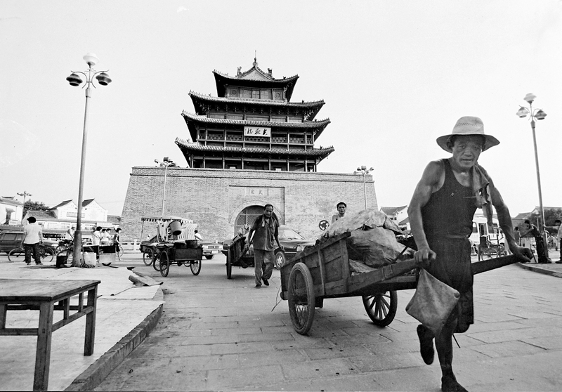 19、《古樓下的板車》1996年8月,聊城市區光岳樓前,兩位男子拉著板車依次前行。在他們身后,小轎車慢慢行駛著。在上世紀90年代,進城的商販運送貨物大多采用這樣的板車。(林虎).JPG